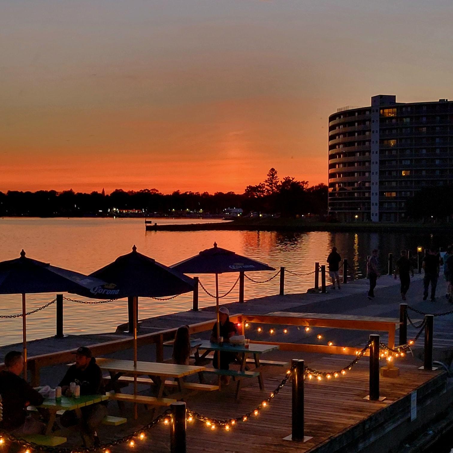 people sitting at tables on a dock at sunset.