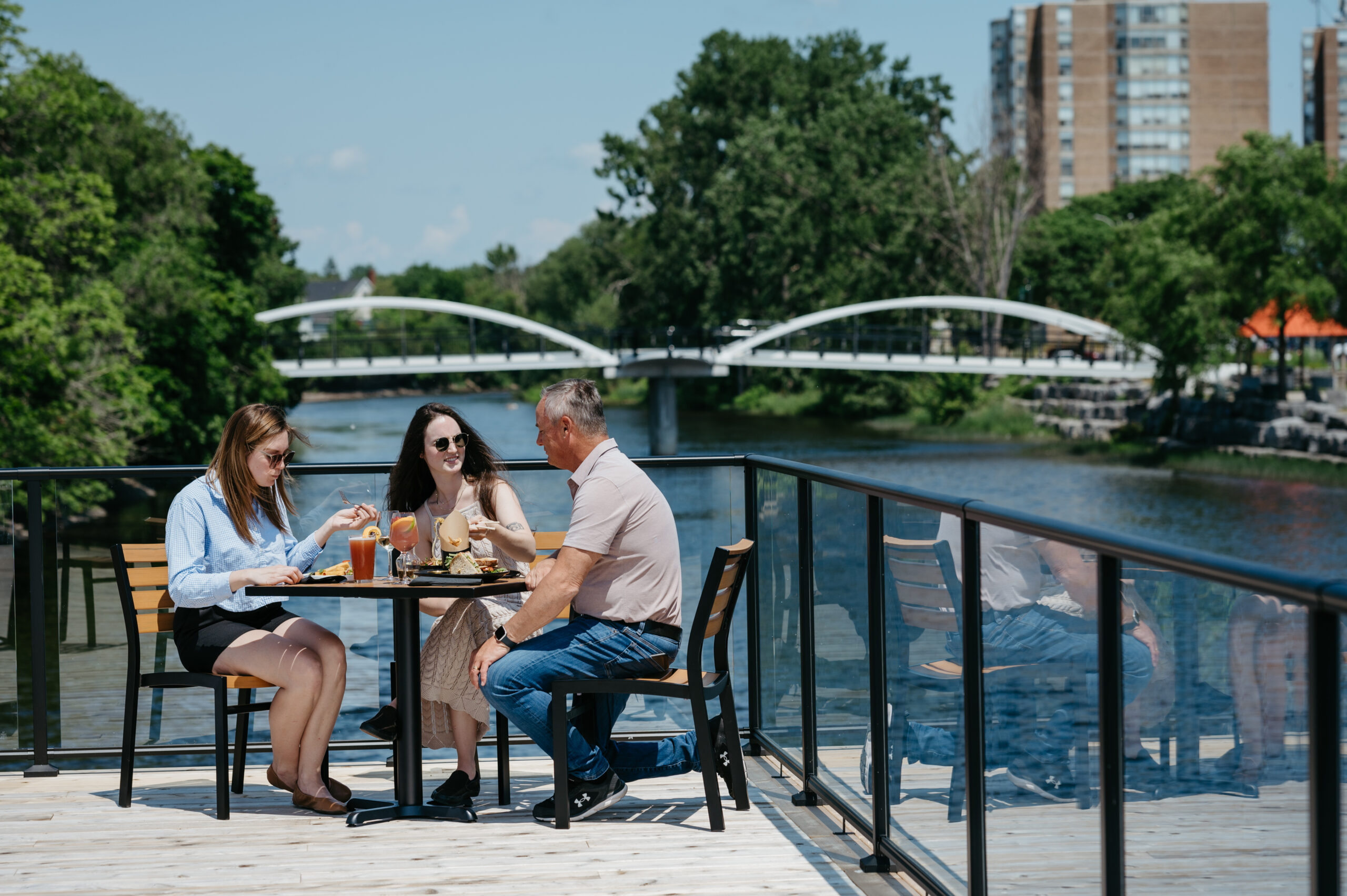 three people sitting on a patio table overlooking a river, a pedestrian crossing bridge in the background