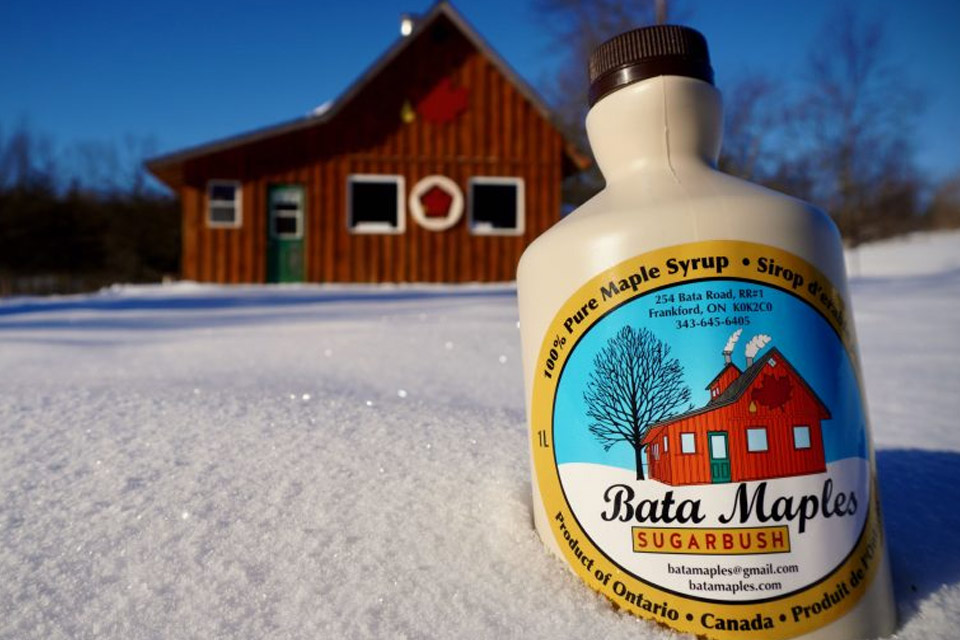 a bottle of maple syrup sits in the snow next to a cabin.
