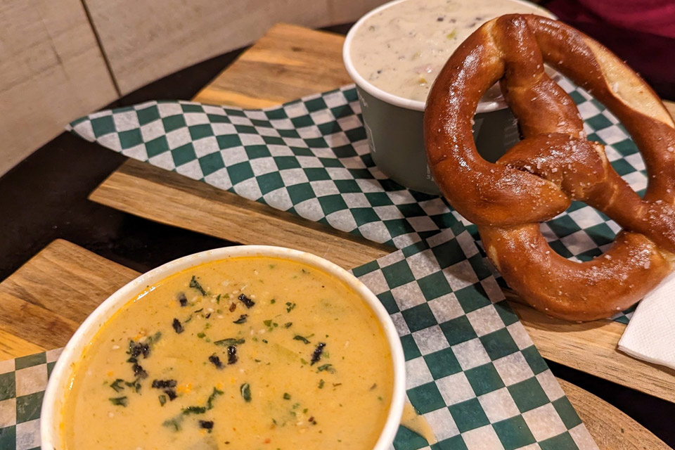 two bowls of soup and a pretzel on a wooden tray.