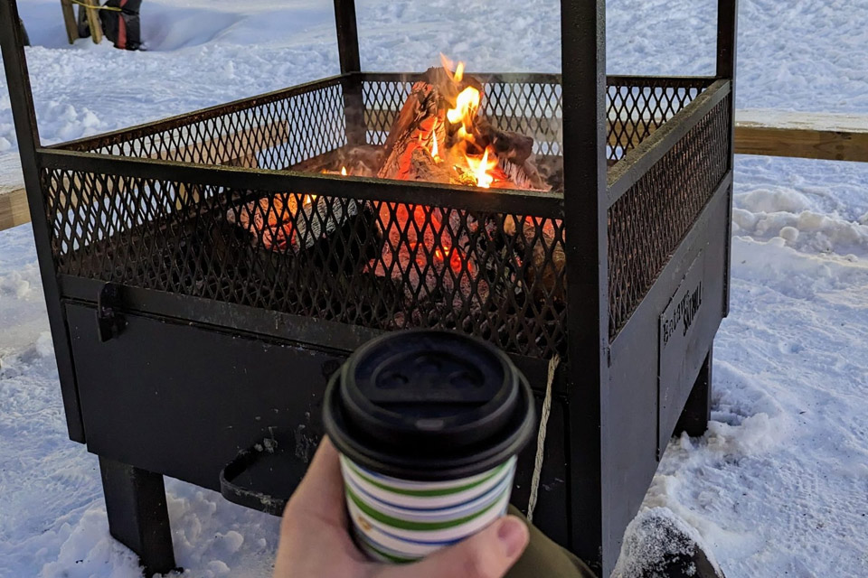 a person holding a coffee cup next to a fire pit in the snow.