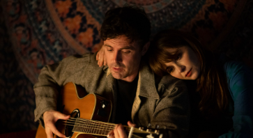 Photo of lead actor playing acoustic guitar with actress leaning on his shoulder in dimly lit room.