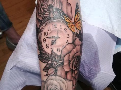 Photo of an arm with clock tattoo surrounded by roses and a butterfly.