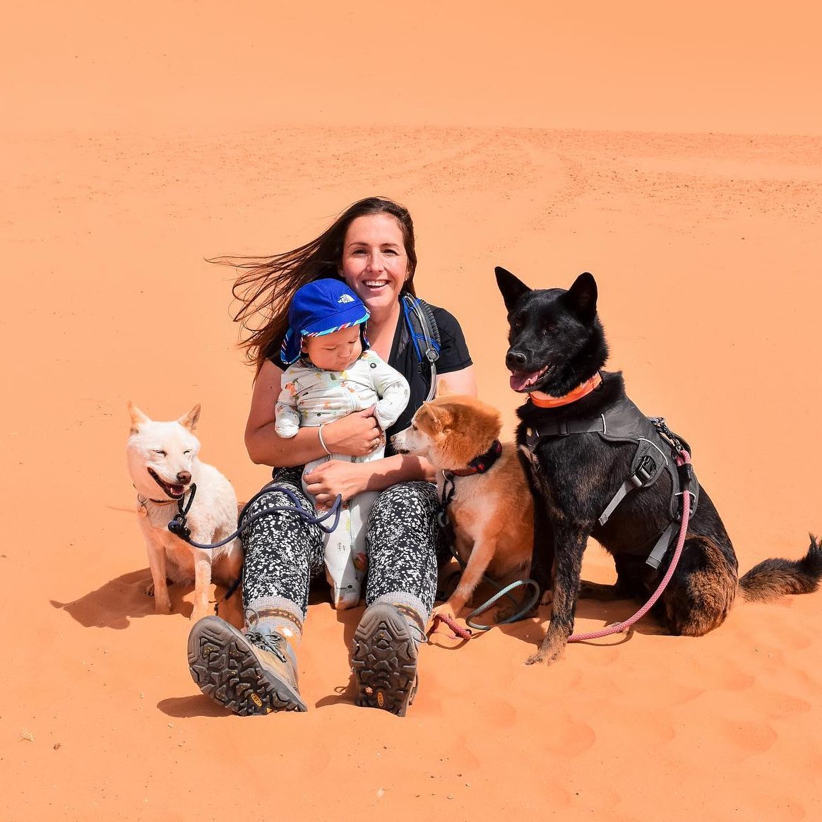 woman holding a baby with three dogs beside them sitting on a pink sand dune