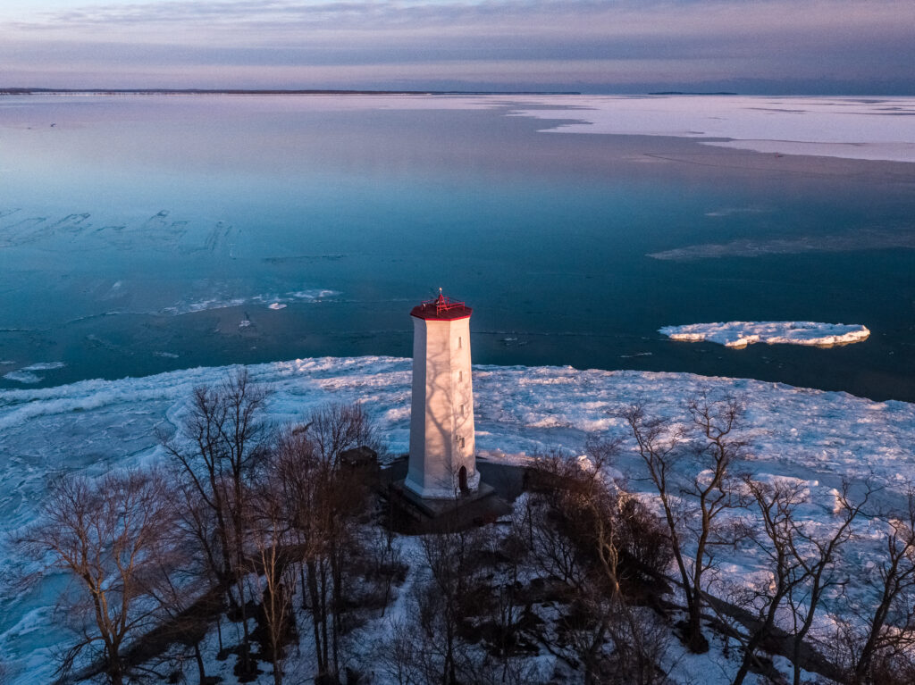 aerial photo of a lighthouse next to open water, snow is on the ground and there is ice in the lake