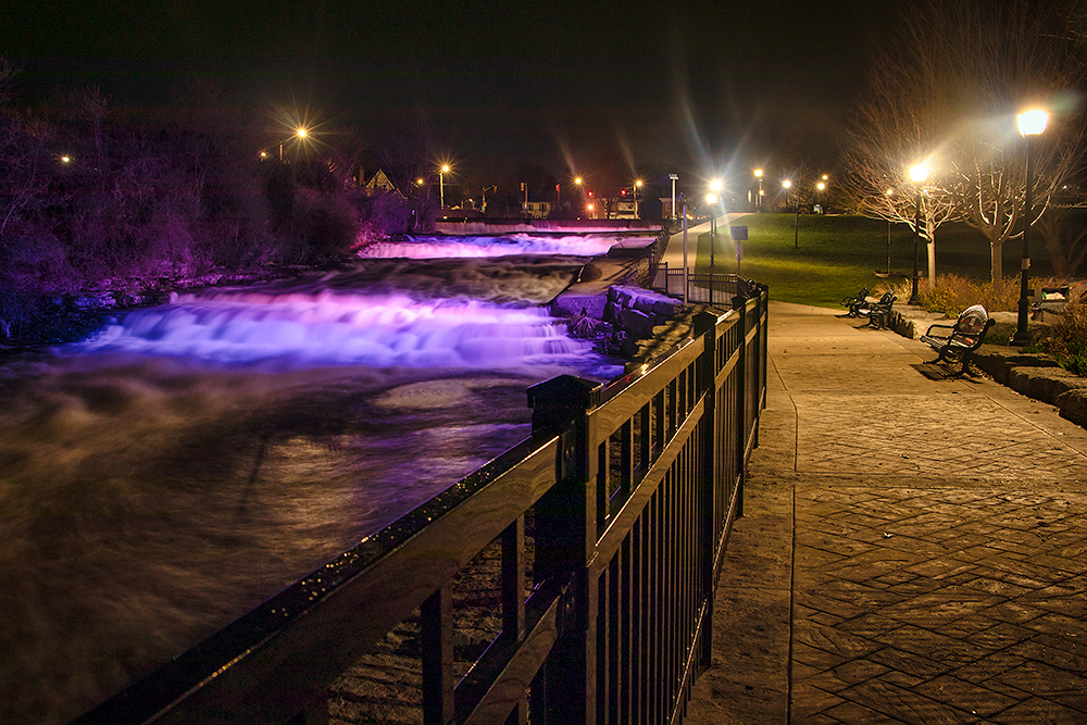 a trail alongside a short waterfall at night that is lit up with purple and pink lights