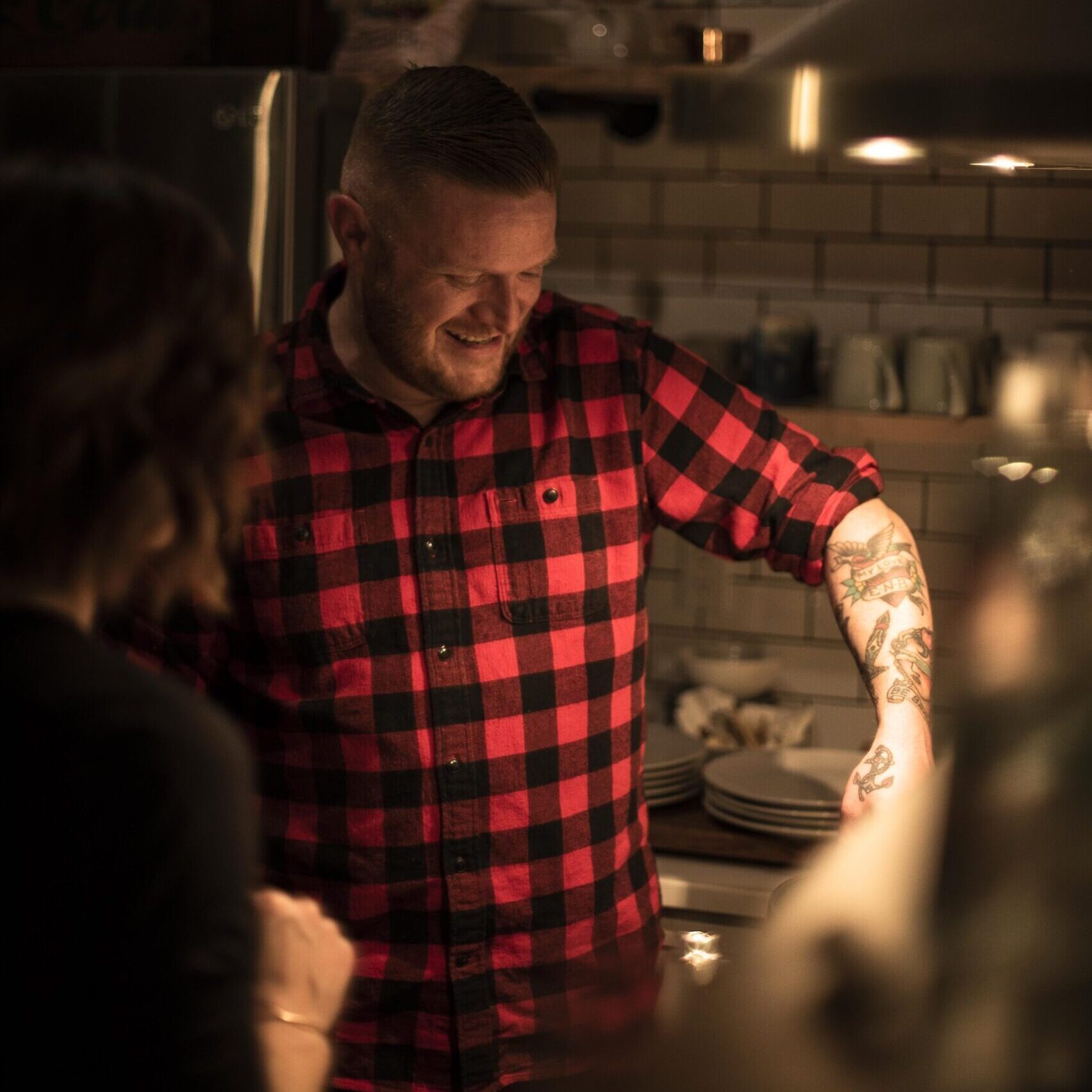 man in a buffalo check plaid shirt with lots of tattoos cooking over a stove in low light