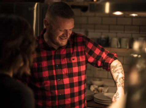 man in a buffalo check plaid shirt with lots of tattoos cooking over a stove in low light