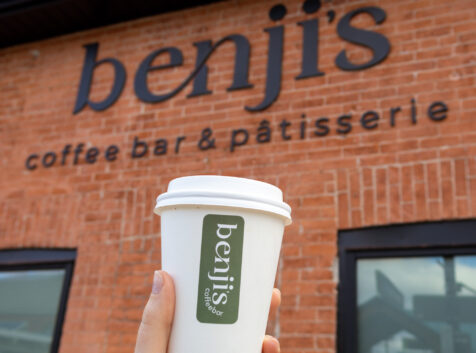 a hand holding up a coffee cup in front of an exterior brick wall with a sign that reads Benji's coffee bar and patisserie
