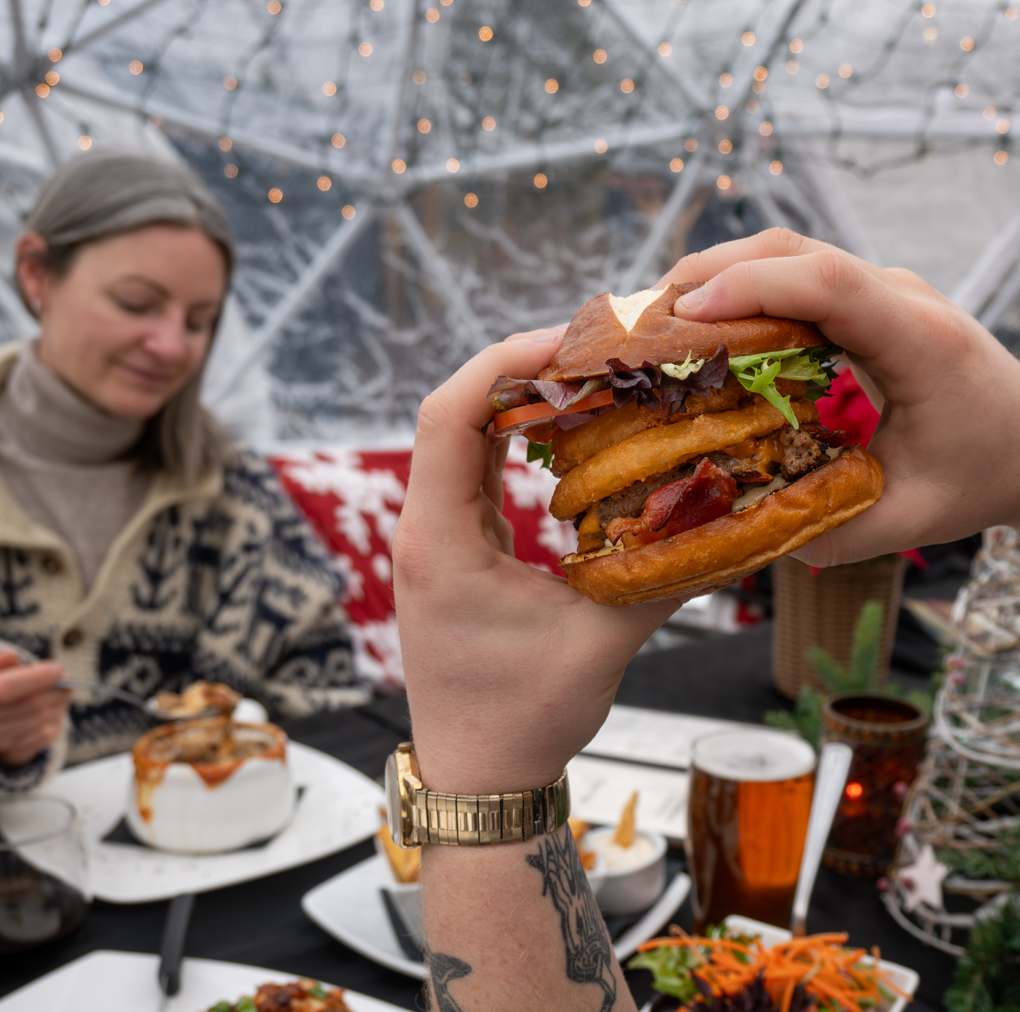 a hand holding up a burger while dining in a snowglobe tent outside in the winter