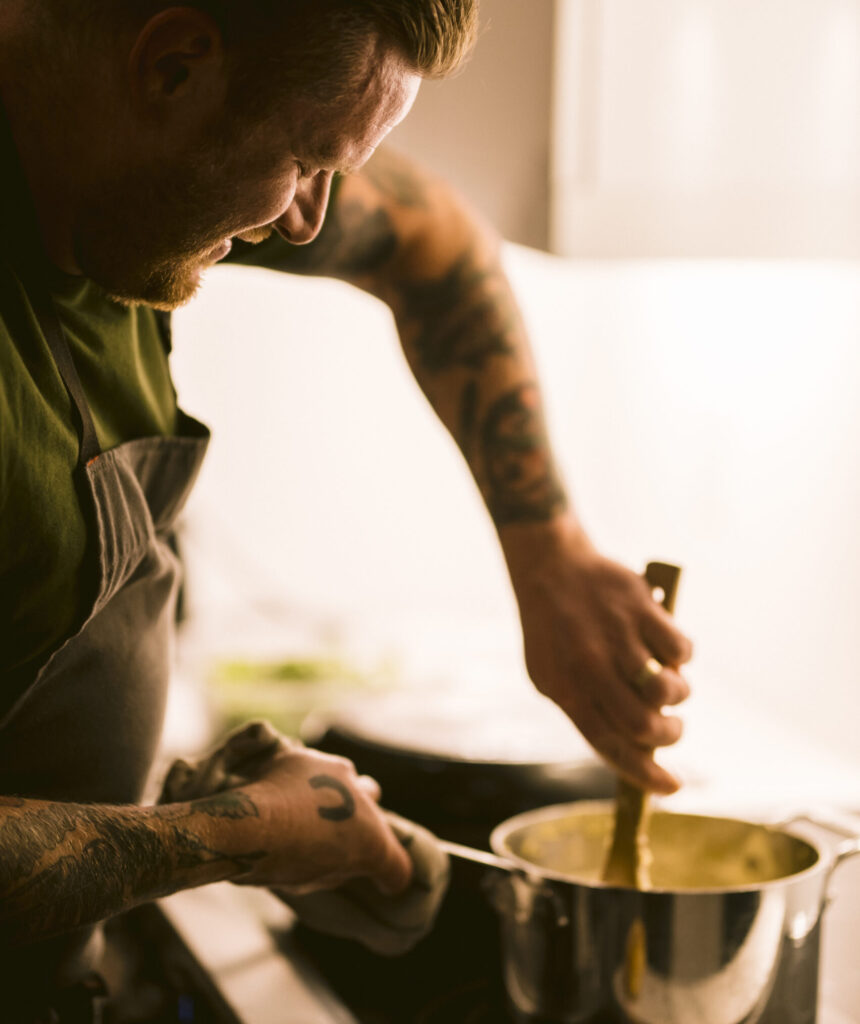 a chef (man with short hair and lots of tattoos) stirring something in a pot over a stove