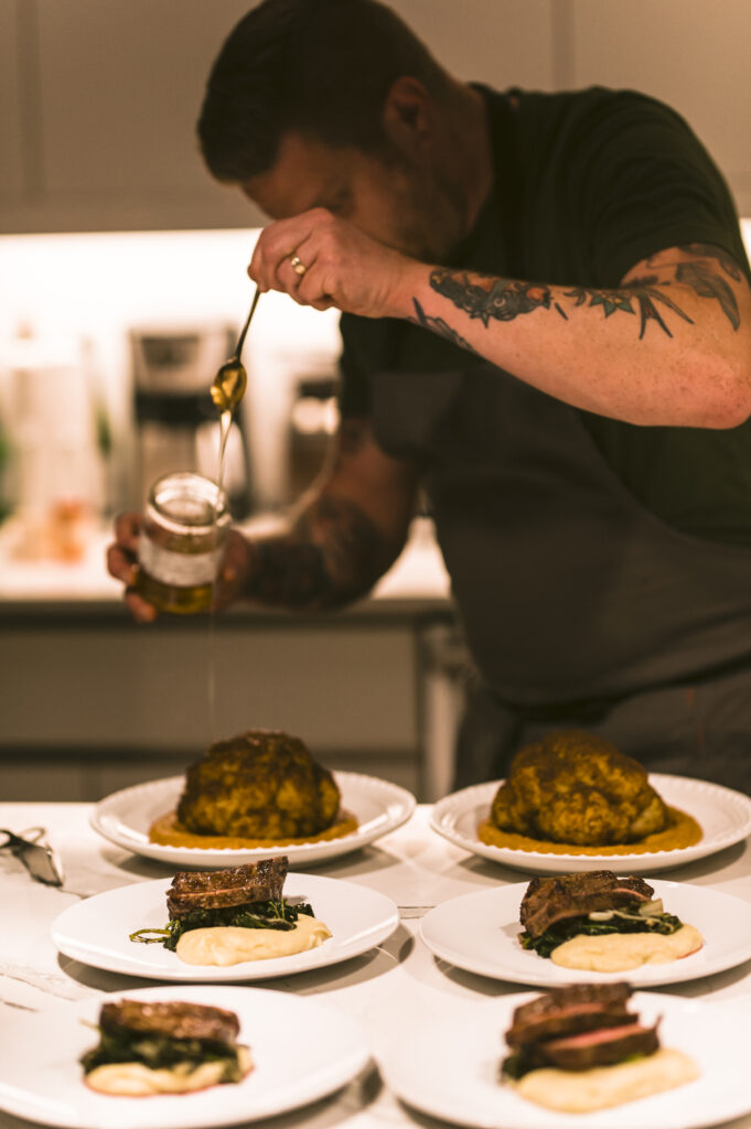 a chef (male, short hair, tattoos on arm), putting finishing garnishes on a few dishes on a counter
