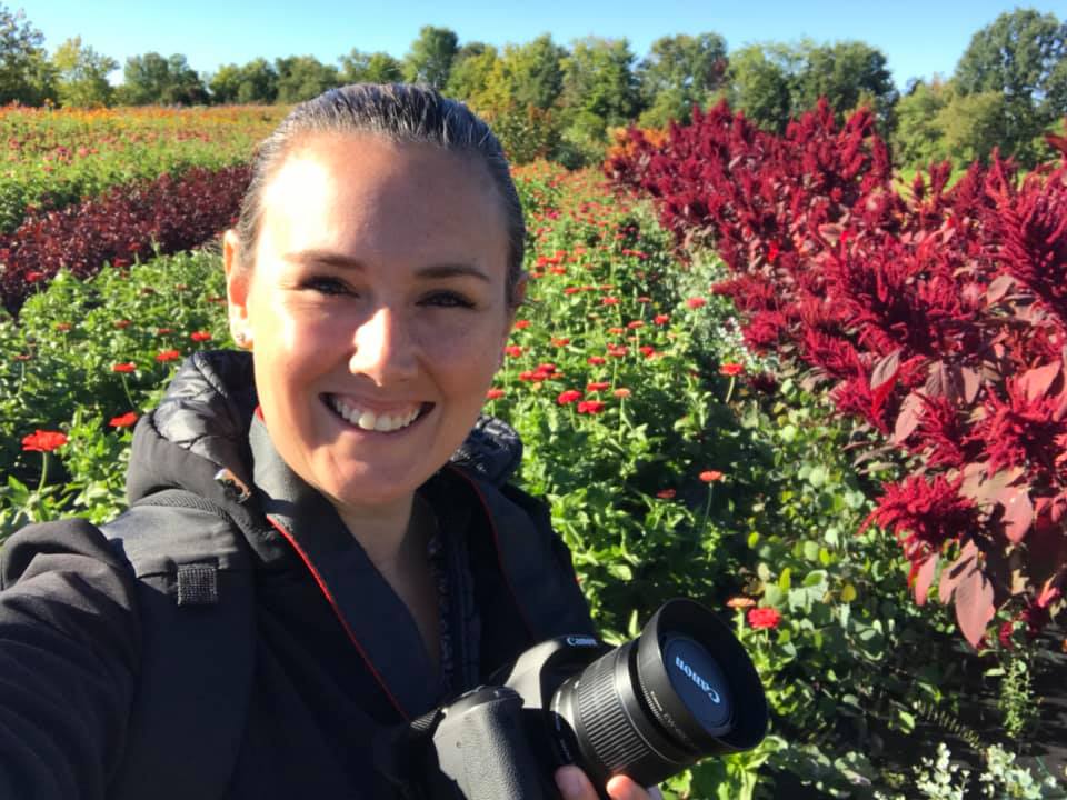 white woman with brown hair taking a selfie while holding a camera in a flower field