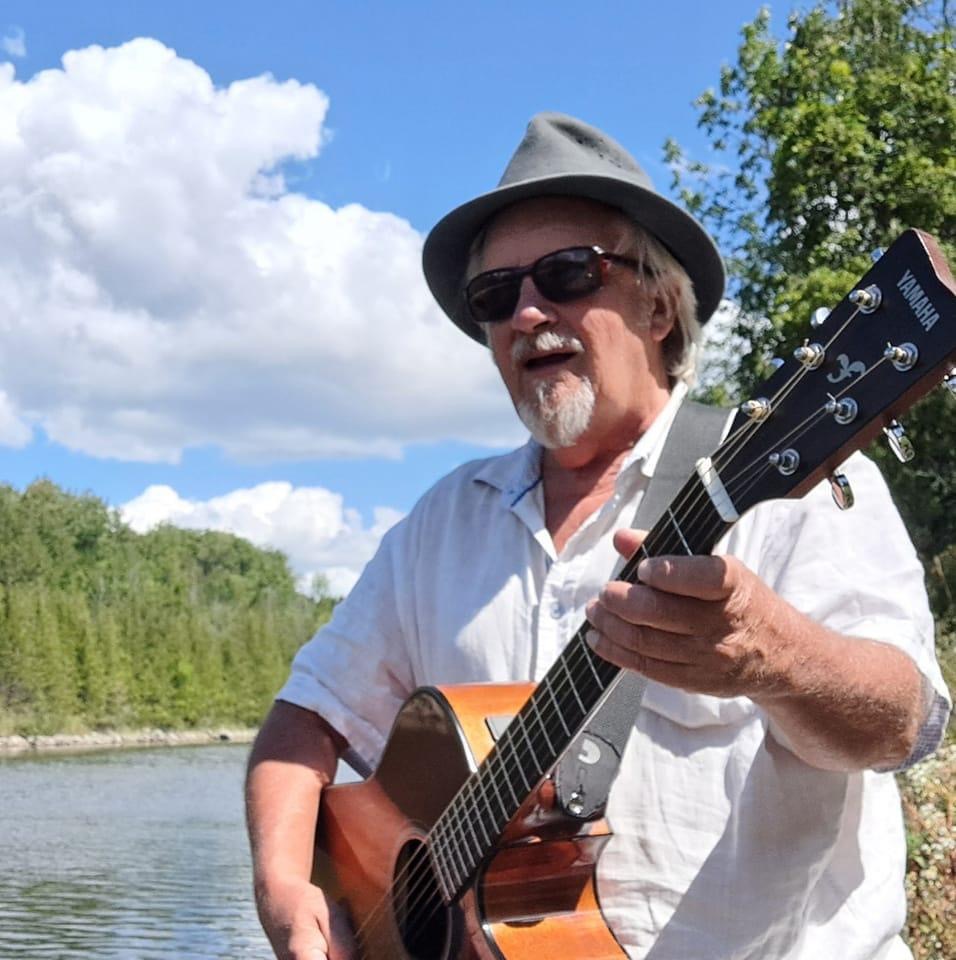 Photo of musician Rick Zimmerman with guitar beside a river.