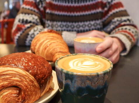 two cappuccinos with latte art and croissants, a person in the background holding their drink