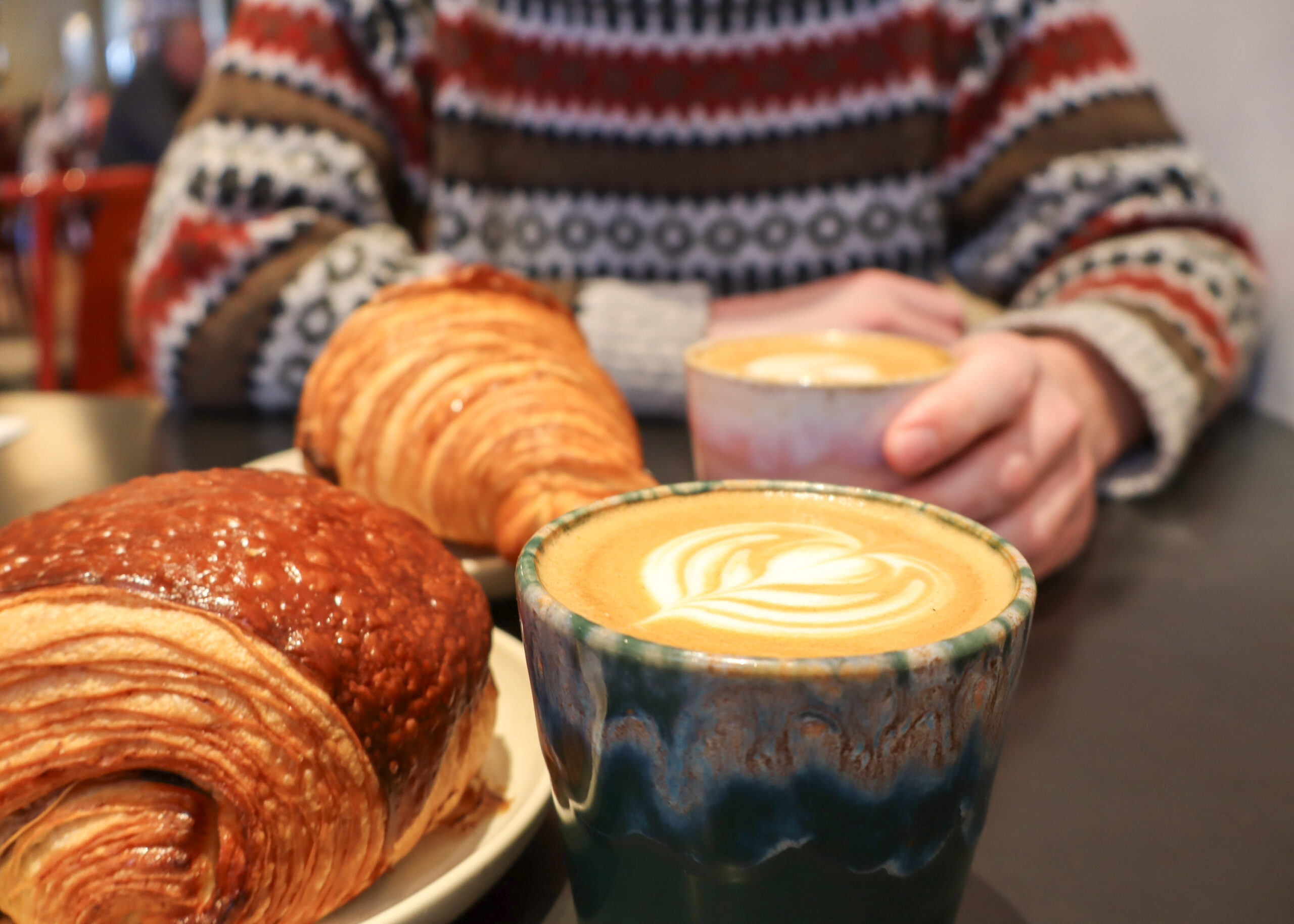 two cappuccinos with latte art and croissants, a person in the background holding their drink