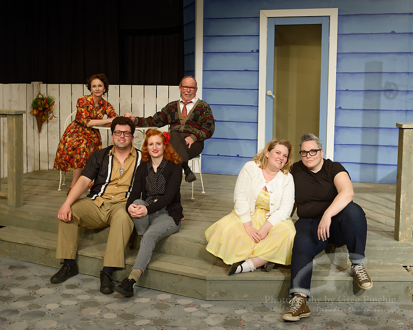 six cast members of a play sit on the steps of a set wearing 1950s attire