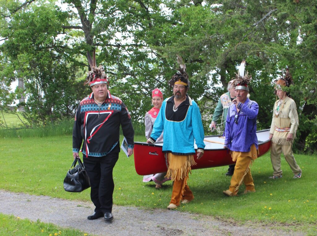 A group of Mohawk people wearing ribbon shirts and other regalia holding a canoe and walking on grass away from the water for the Mohawk Landing celebration.