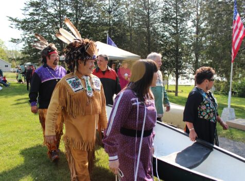 a group of Mohawk people dressed in regalia holding a canoe and walking on grass towards the water for the Mohawk Landing celebration.