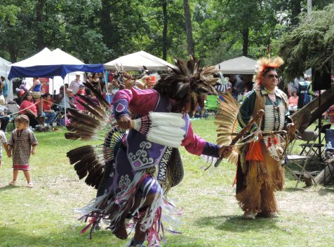 Two Indigenous people dancing while dressed in traditional regalia at a Pow Wow