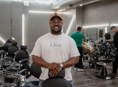 Kam, owner of revive studios, poses for a photo leaning on one of the barber chairs with two employees working in the background